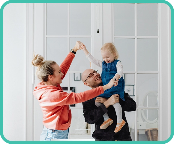 Two adults lift and play with a young girl in a bright room with large glass doors in the background.